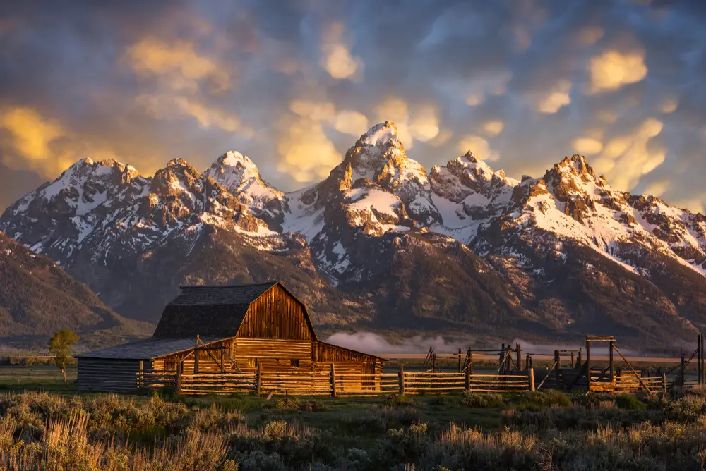 Teton range, a must-visit place in Wyoming, pictured at sunset on a clear day with cottonball-like clouds overhead of a barn