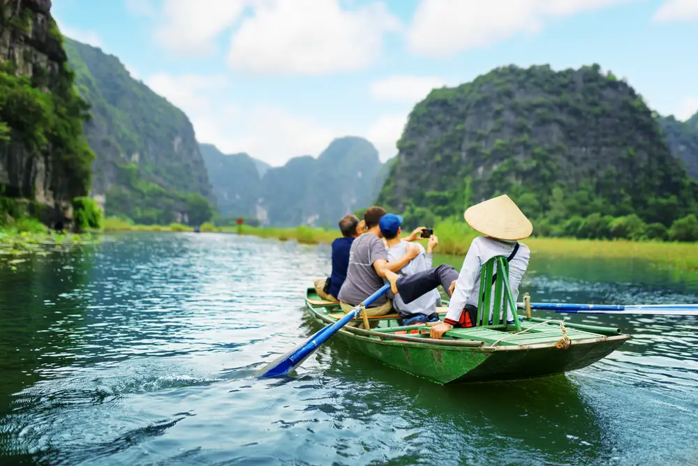 Tourists riding along in a small green rowboat pictured from the back with a pointy-hat guide in a white shirt guiding them down the Ngo Dong River, included for a piece titled Is Vietnam Safe to Visit