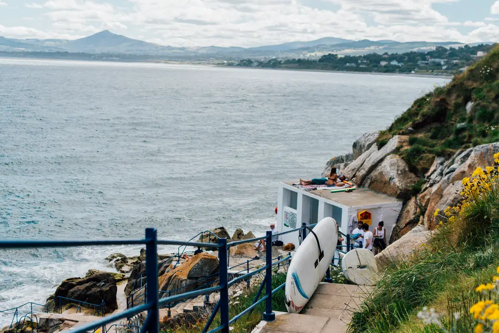People on a cement boardwalk in Dalkey, one of the best places to stay in Dublin, pictured on a cloudy day with a surfboard on the steps overlooking the cliffs