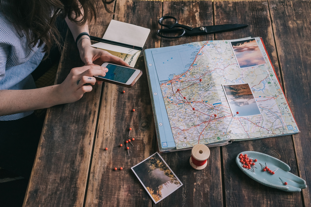 Woman uses her smartphone with a map and travel documents on a table to plan attractions to visit and stops along the way for her trip