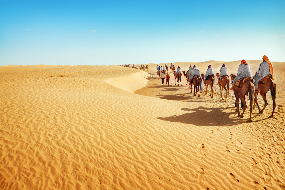 Ομάδα ανδρών σε ένα τροχόσπιτο με καμήλα διασχίζοντας την έρημο Σαχάρα για να δείξει την καλύτερη στιγμή για να επισκεφθείτε την Τυνησία τον Απρίλιο έως τον Ιούνιο