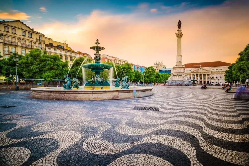 Η πλατεία Rossio απεικονίζεται με το μοναδικό κυματιστό δάπεδο με πλακάκια με μοτίβο ζέβρας, ένα σιντριβάνι και ένα τεράστιο άγαλμα με θέα στην πλατεία για ένα κομμάτι με τίτλο Where to Stay in Lisbon