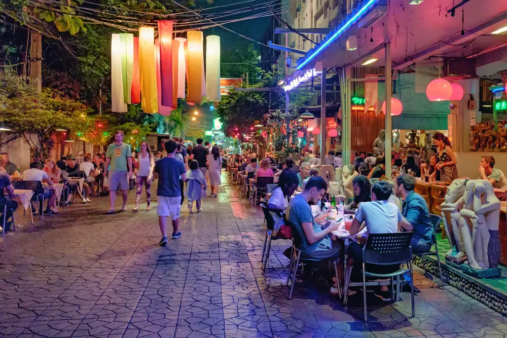 Locals and tourists sitting on chairs in an outside market in Rambuttri Alley at night, close to Khaosan Road to illustrate that Bangkok is safe to visit