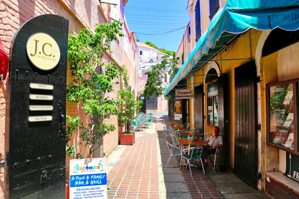 For a guide to whether or not the Virgin Islands are safe to visit, a photo of a jewelry store with a man sitting outside under a blue sky with a brick walking path down the narrow and well-lit alleyway