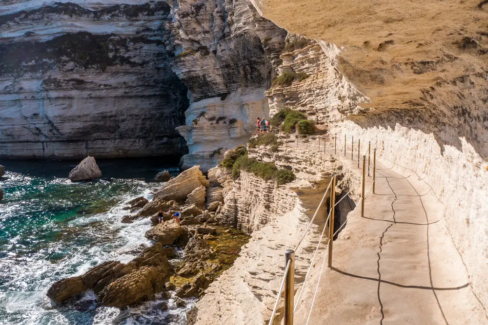 Picturesque and serene scene with people climbing the rocks below the stone walking path next to a rope and metal handrail, seen during the best time to visit the Mediterranean