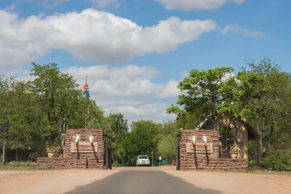 Neat view of the tusk-lined Oilfants Restcamp Gate with a car driving through it below a cloud-covered sky for a guide to whether or not Kruger National Park is safe to visit