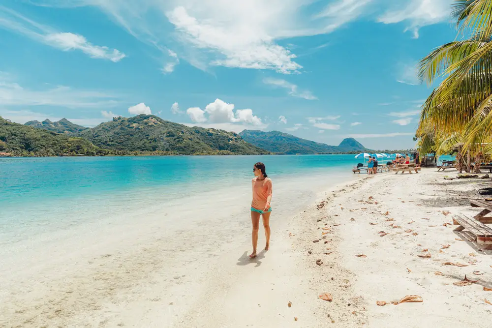 Woman in jean shorts and a peach-colored shirt walks along the white sand beach in Huahine, French Polynesia, to illustrate that Tahiti is perfectly safe to visit when the right precautions are taken