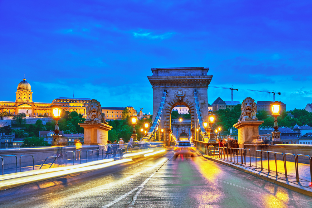 Low-shutter image showing cars driving by the camera with a dark blue sky overhead in front of the Royal Castle and Czechenyi Chain Bridge