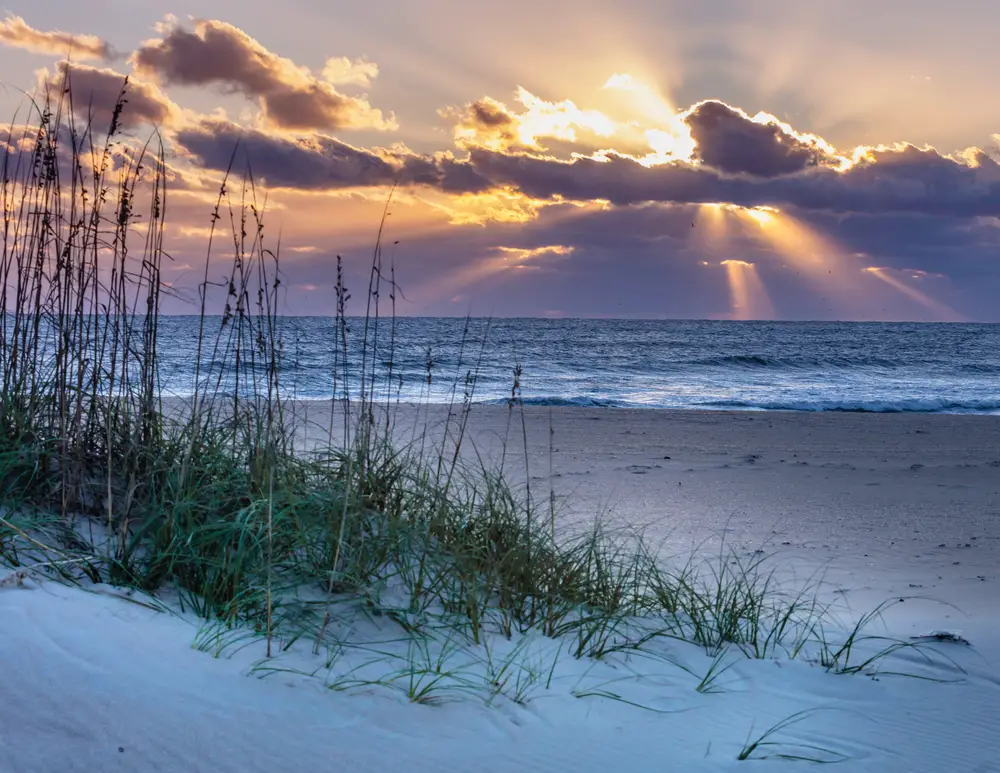 Concept of the best couples vacations in the US with imagery of Ocracoke Island featuring seagrass and sun rays peeking through the clouds at sunset