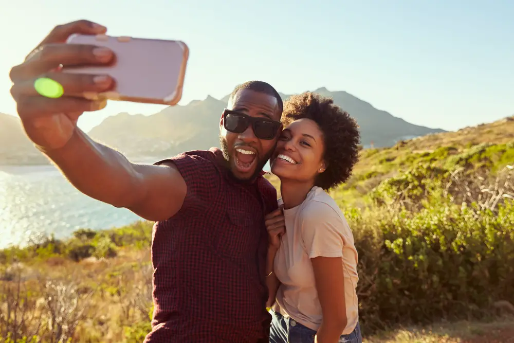 Young African American couple smiling and taking a selfie outdoors with mountains in the distance at one of the best couples vacations in the US