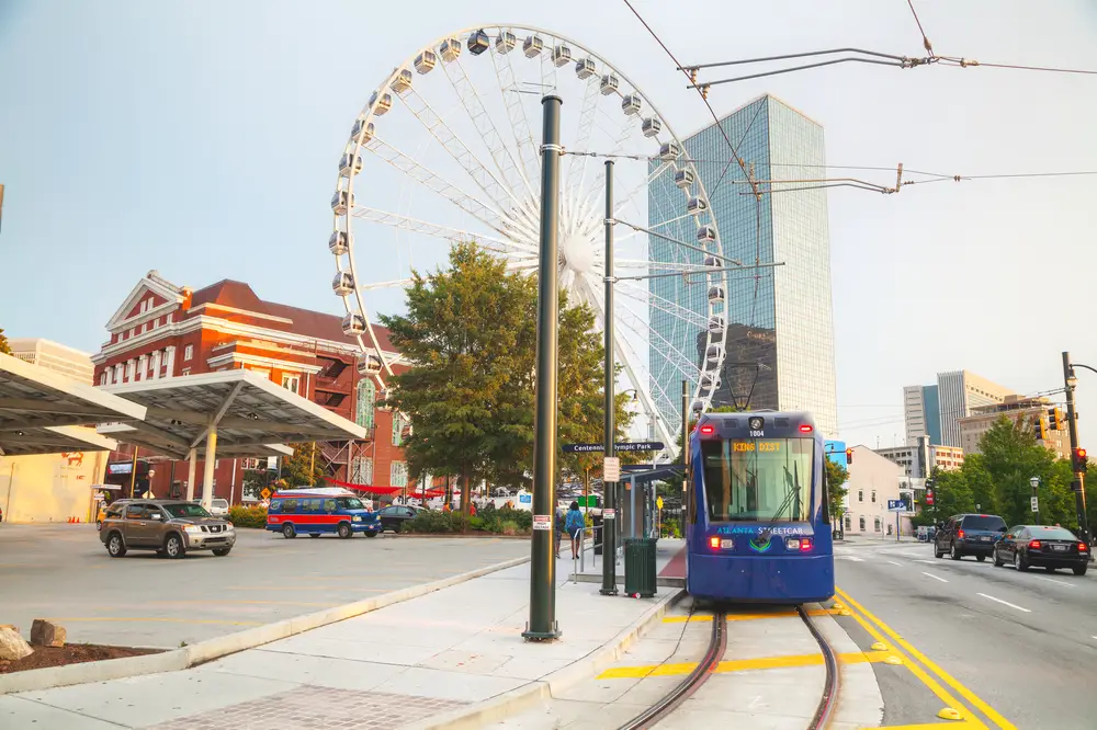 Photo of a Ferris Wheel pictured outside of the downtown area with a blue light rail train on the street next to a gas station