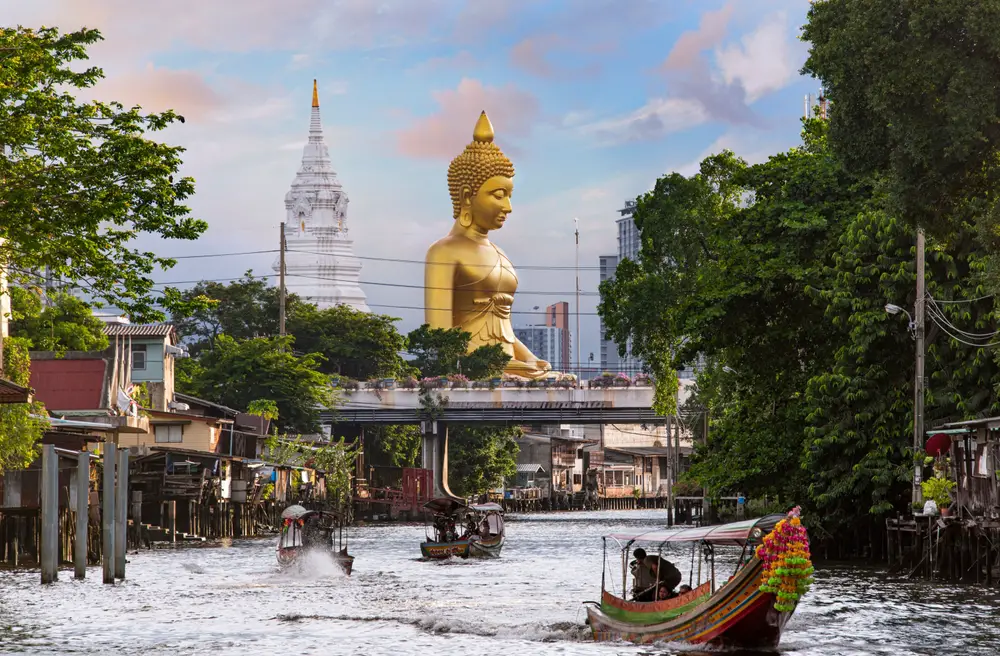 Photo of a giant golden statue pictured towering high above a canal with boats on it in Bangkok