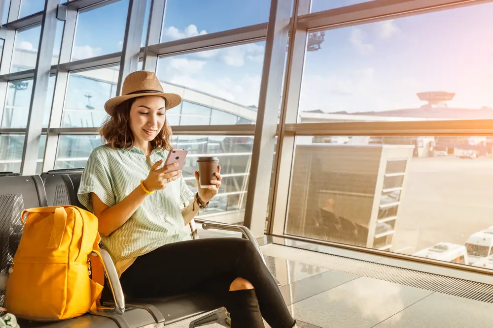 Νεαρή γυναίκα που κάθεται σε ένα αεροδρόμιο με τις κίτρινες αποσκευές της δίπλα της χρησιμοποιεί το smartphone της για να επιβεβαιώσει τα στοιχεία της πτήσης της στο Google Flights