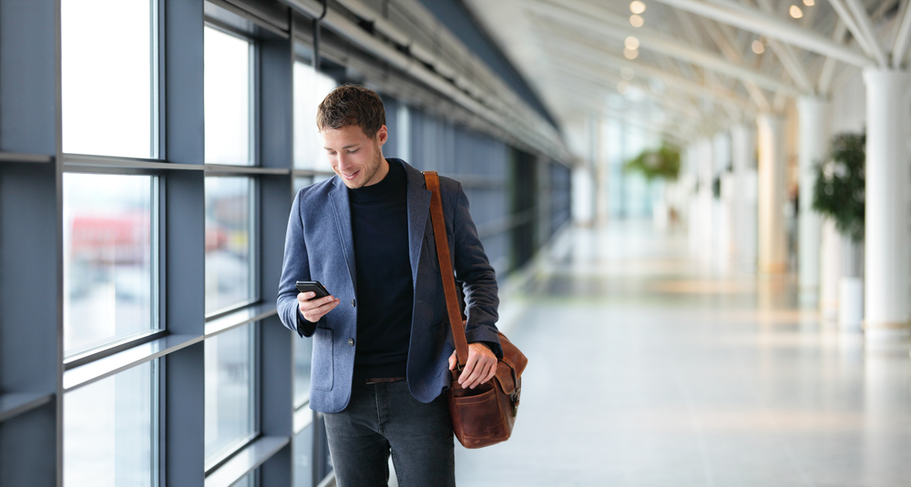 Άνδρας που περπατά μέσα από έναν τερματικό σταθμό αεροδρομίου ρίχνει μια ματιά στο smartphone του αφού έμαθε πώς να χρησιμοποιεί τις Πτήσεις Google για να κάνει κράτηση και να αναζητά πτήσεις πολλών πόλεων καθώς ταξιδεύει