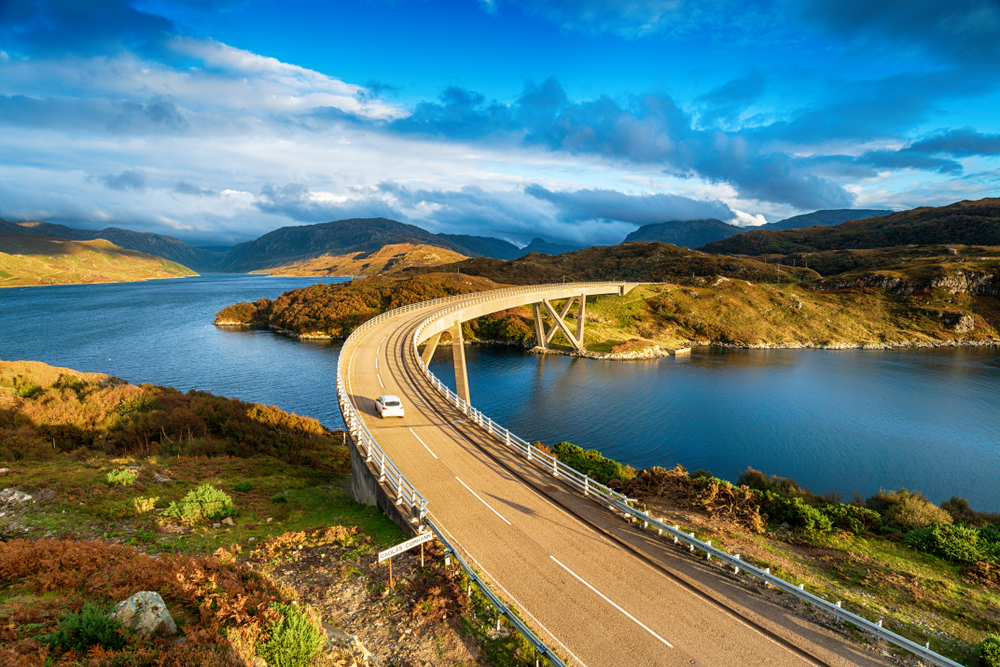 Για μια εικόνα για έναν οδηγό για το μέσο κόστος ταξιδιού στη Σκωτία, μια φωτογραφία ενός λευκού αυτοκινήτου που οδηγεί στην αριστερή πλευρά του δρόμου πάνω από τη γέφυρα Kylesku