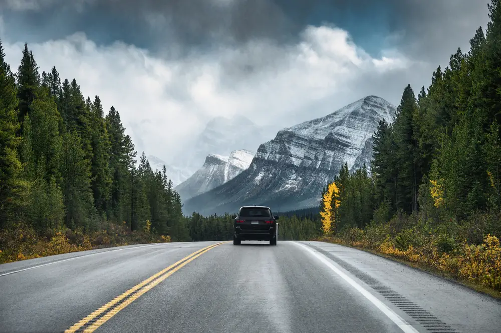 Dark SUV driving down a highway with mountains in front and clouds overhead for an article asking can you take rental cars out of state?