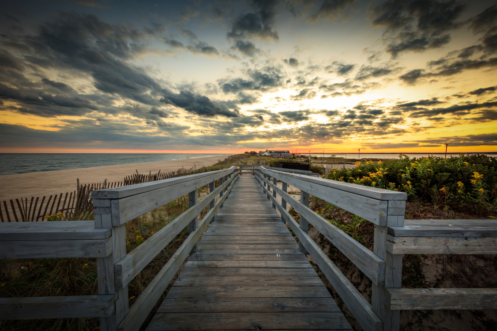 For a guide to the best and worst times to visit the Hamptons, a long wooden boardwalk leading to the ocean is seen at dusk