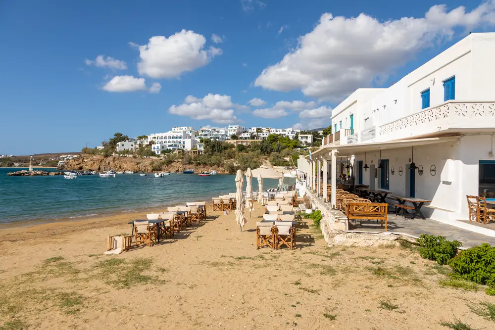 Small white buildings pictured in Piso Livadi, one of the best places to stay when in Paros, pictured with tan sand by the ocean