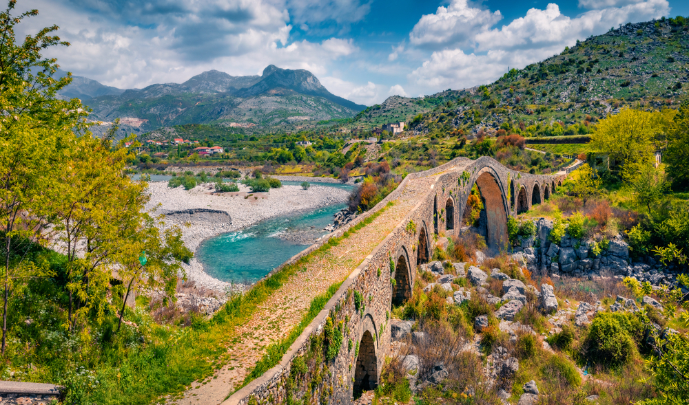 Old Mes Bridge in Albania during summertime with clouds in the sky and blue water with mountains in the distance shows Albania to be one of the best cheap places to travel to in the world