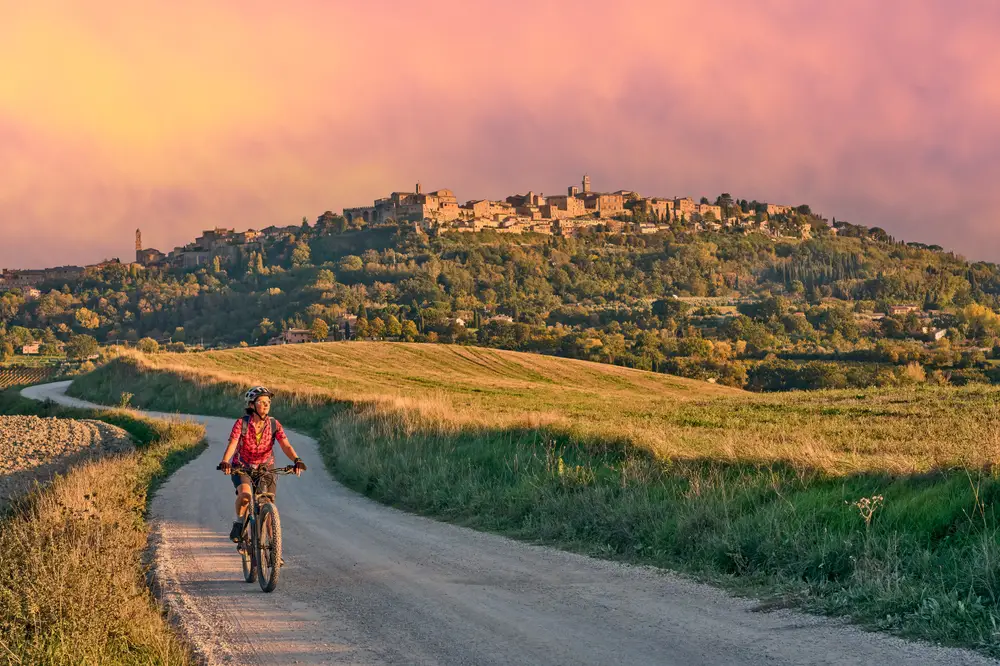 Woman riding a bike in the middle of a late-summer evening between olive trees in the Ghianti Area with Montepulciano in the background, pictured for a guide on the average European trip cost