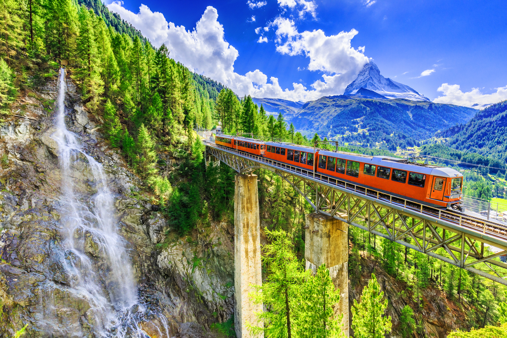 Φωτογραφία του κόκκινου τρένου που κάνει το δρόμο του πάνω από τη γέφυρα πάνω από την κοιλάδα στο Zermatt για έναν οδηγό για το μέσο κόστος ταξιδιού στην Ελβετία