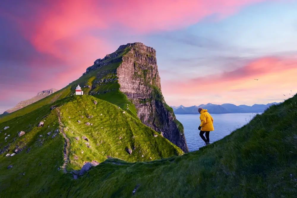Guy in a yellow coat pictured walking along the cliffside in Kalsoy Island for a guide to whether or not the Faroe Islands are safe to visit