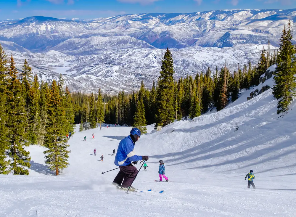 People skiing down the snow and an area of green trees is sitting at the bottom with snowy mountains in background. 