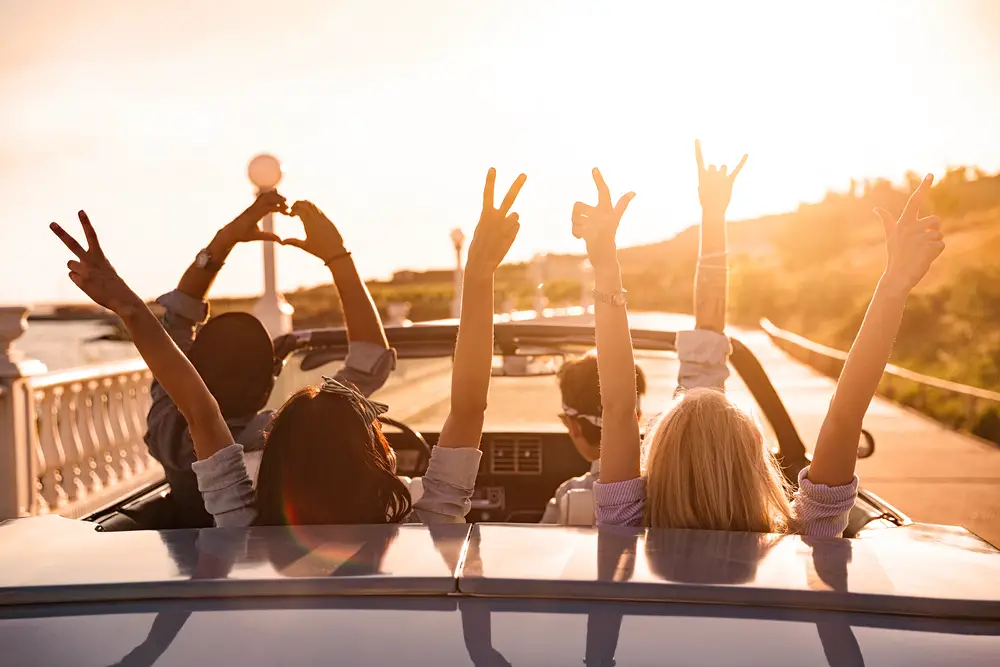A group of friends takes off on a trip in a convertible with hands in the air at sunset for a frequently asked questions section on the best trips for groups of friends