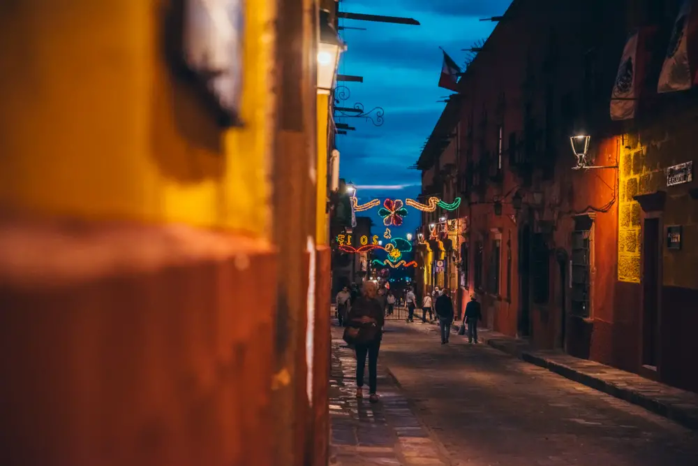 Several people walking down an alley illuminated the street lamps at dusk. 