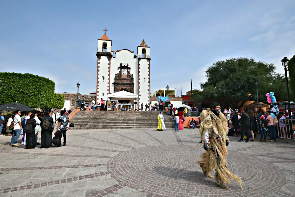 A local man wearing traditional clothing passing a the front of a church, while tourists are busy scanning the place. 