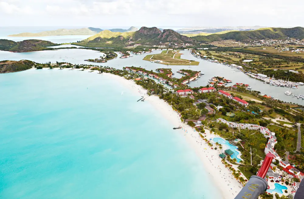Aerial view on a long shoreline with white sand and clear water, with resorts and hotels and boats. 