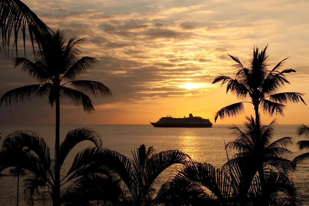 Hawaii cruise ship sailing at sunset with palm trees in the foreground for a guide listing the ways for traveling between the islands in Hawaii