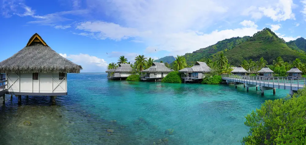 Bungalow houses erected few feet above tropical waters with a foot bridge and a green mountain. 