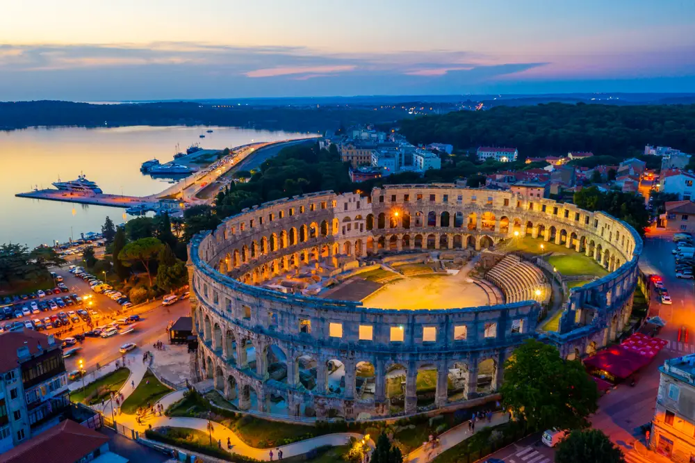 A historical amphitheater preserved through time, seats in a city surrounded by modern structures, and a calm bay in can be seen in background during sunset. 