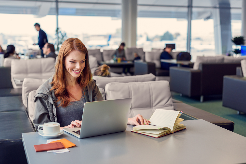 Smiling businesswoman in an airport lounge uses her laptop with a small crowd of others in the background to show if Priority Pass lounges are usually crowded