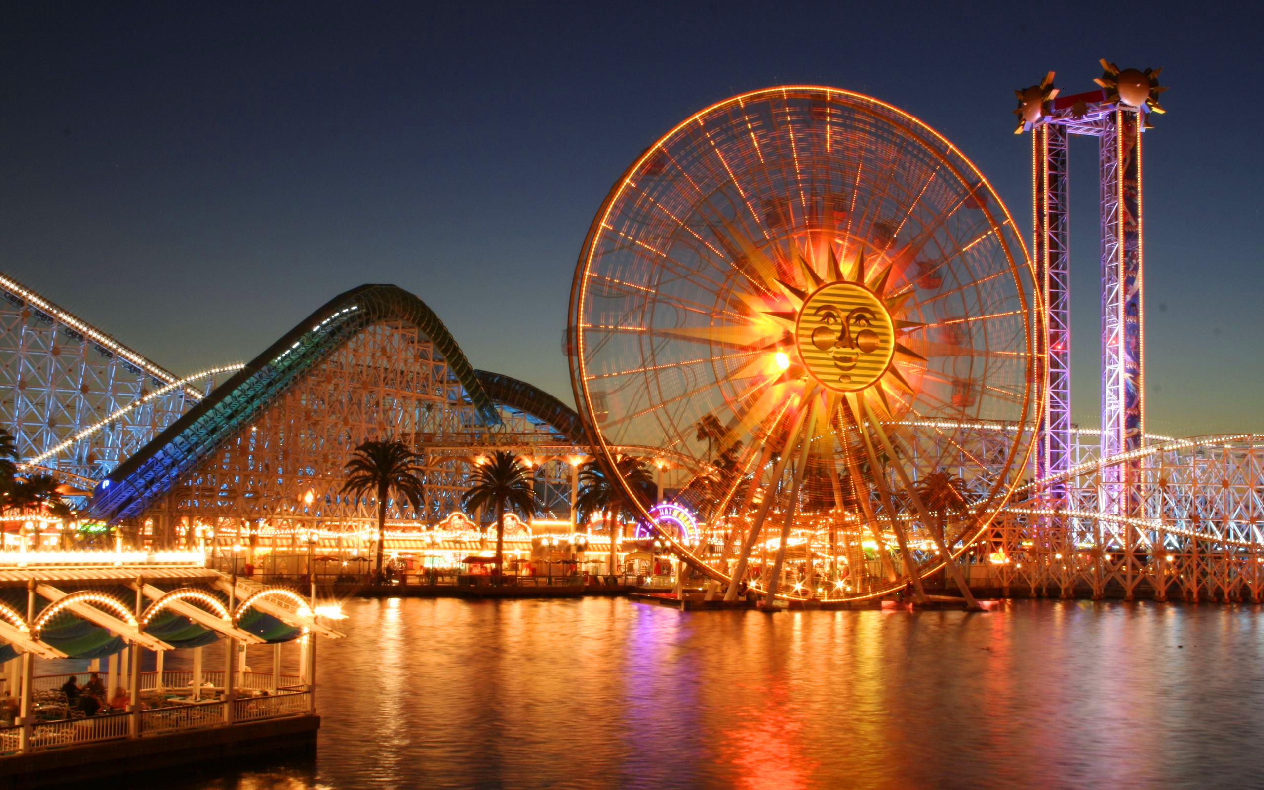 A gigantic Ferris Wheel with a sun image in the middle, and other rides in an amusement during dusk. 
