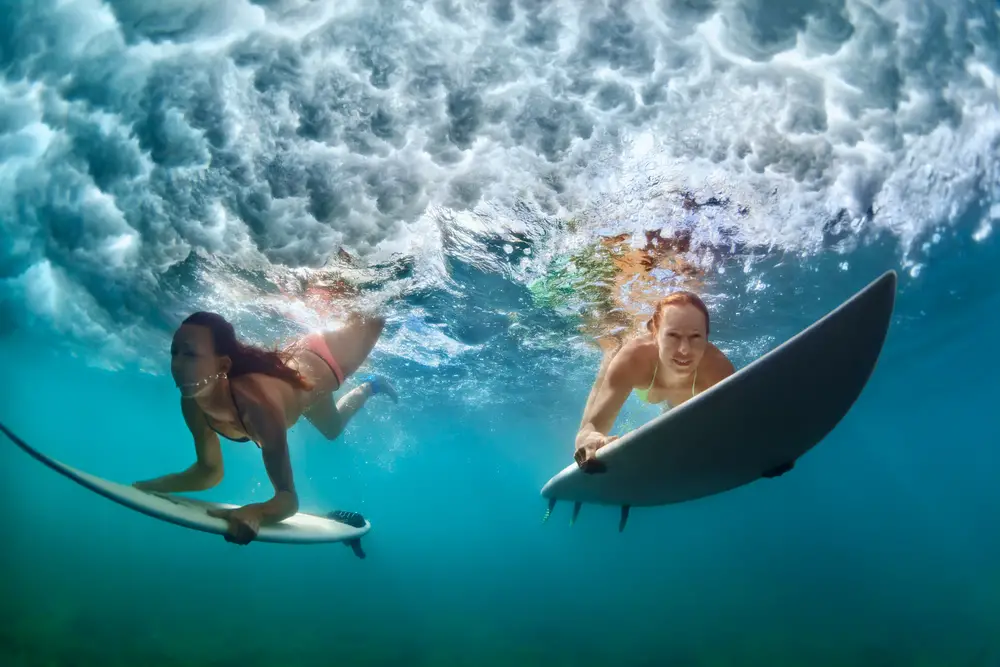 Two women in their swimwear diving down the surface of the water with their surf boards. 