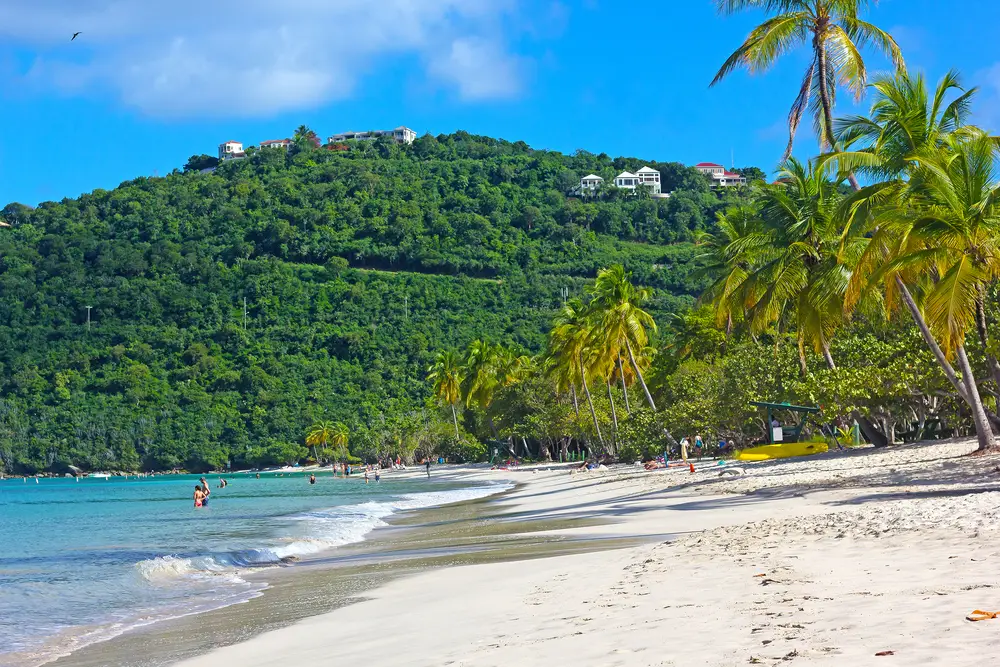 Gorgeous white-sand beach of Magens Bay pictured on a nice day with a few waves lapping the beige sand and a few tourists in the water, below towering vegetation-covered hills pictured for a guide to whether or not St. Thomas is safe to visit