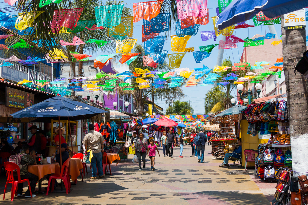 Image of people shopping at the Plaza Santa Cecilia pictured for a guide titled Is Tijuana Safe with Mexican flags above the open-air market, with its many shoppers and lots of shops and restaurants