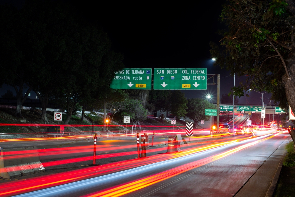 Η Τιχουάνα Ρίο και τα σύνορα του Σαν Υσίδρο απεικονίζονται σε μια εικόνα μακράς έκθεσης με μια θαμπάδα από τα φώτα του αυτοκινήτου να κάνουν τον δρόμο τους από αριστερά προς τα δεξιά κάτω από τις πράσινες πινακίδες