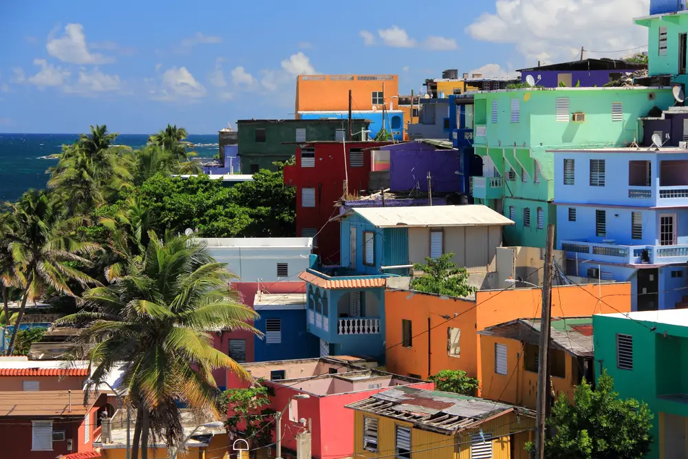 Φωτογραφία σπιτιών στην περιοχή La Perla που απεικονίζεται για έναν οδηγό με τίτλο Is San Juan Safe to Visit με πολλά πολύχρωμα κτίρια στην πλαγιά ενός λόφου με μπλε ουρανό και ωκεανό στο φόντο