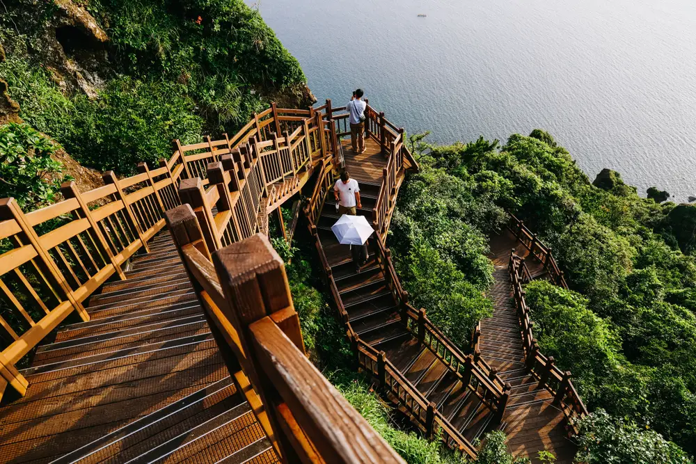 Three people walking on a long stairs on the side of the mountain, and below a calm sea can be seen. 