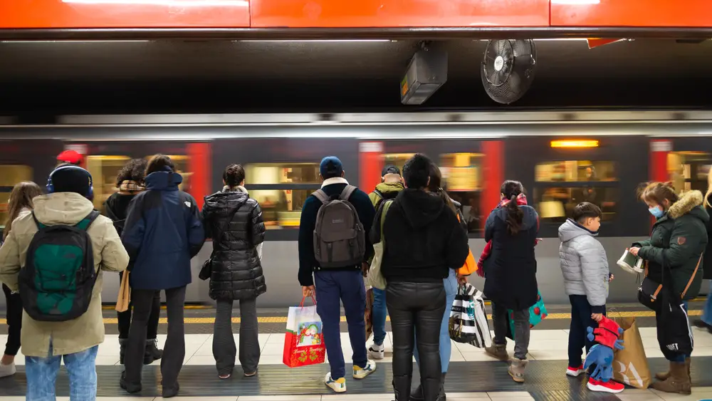 Άνθρωποι που φορούν χειμωνιάτικα μπουφάν στέκονται μπροστά από ένα τρένο που τρέχει περιμένοντας να επιβιβαστούν.