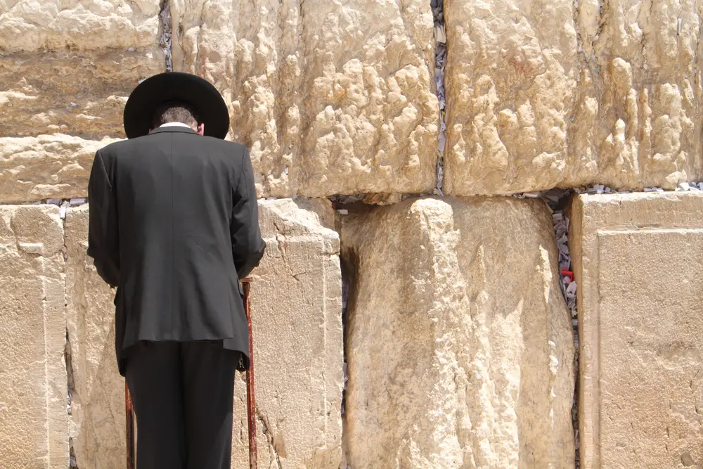 Ένας άνδρας που φορά μια βάρκα και ένα καπέλο φαίνεται να προσεύχεται ενώ κοιτάζει έναν τοίχο από τεράστιους βράχους. 