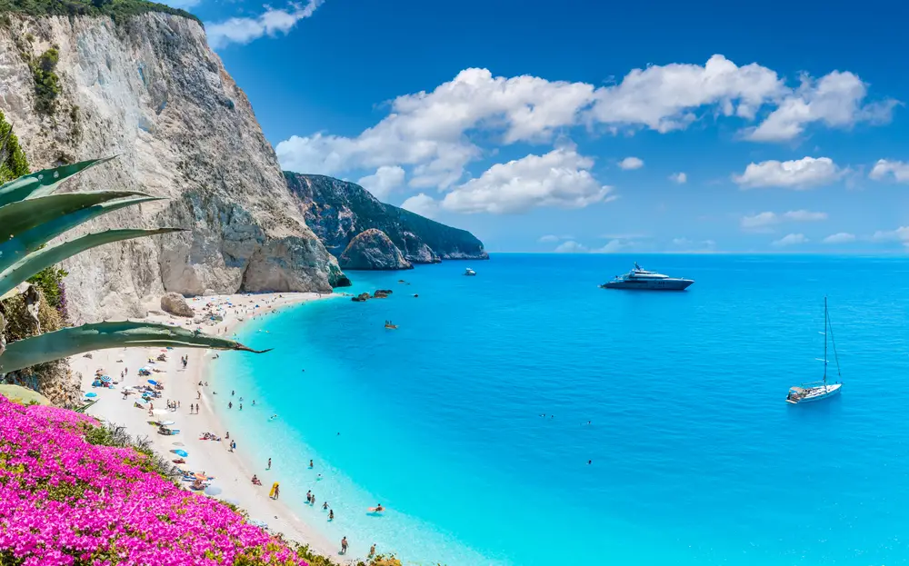 Landscape of Porto Katsiki Beach pictured for a guide titled Is Greece Safe to Visit, with a giant towering cliff on the left and a teal and dark blue beach on the right