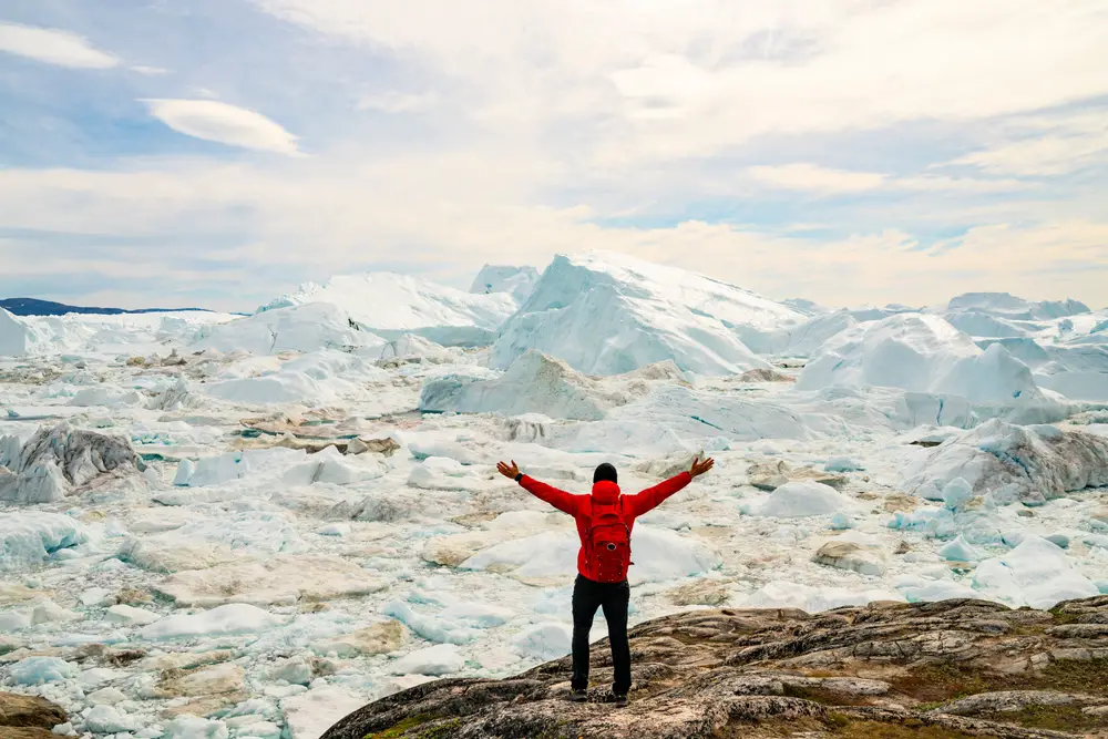 Man in a red coat and black gloves holding his arms above his head while he gazes out over an iceberg field for a guide titled Is Greenland Safe to Visit