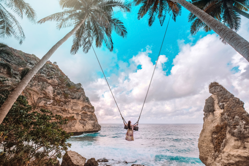 Girl swinging while solo traveling in Bali, Indonesia in front of a beautiful beach with rock formations and palm trees