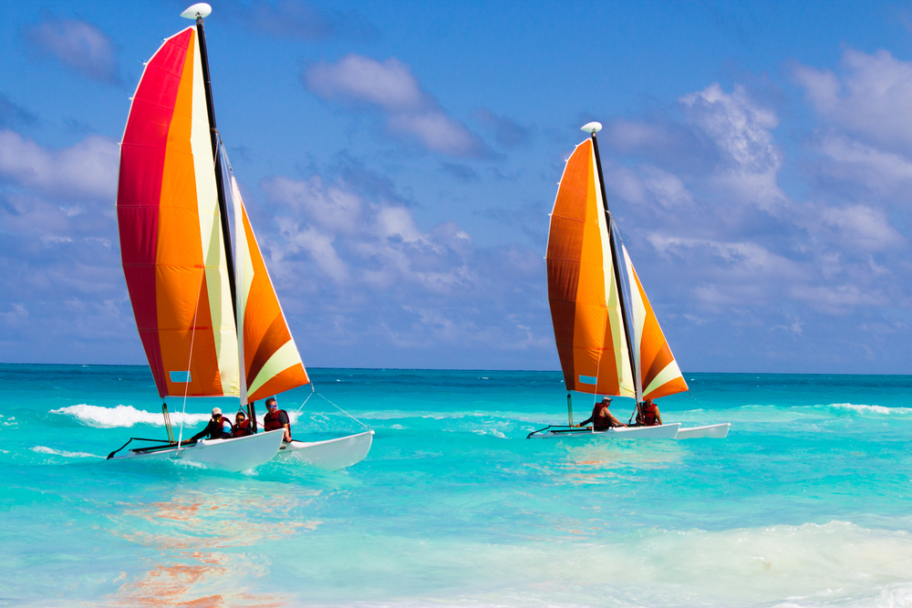 Two small vibrant sailboats with a few passengers nearing the shallow waters of a beach. 