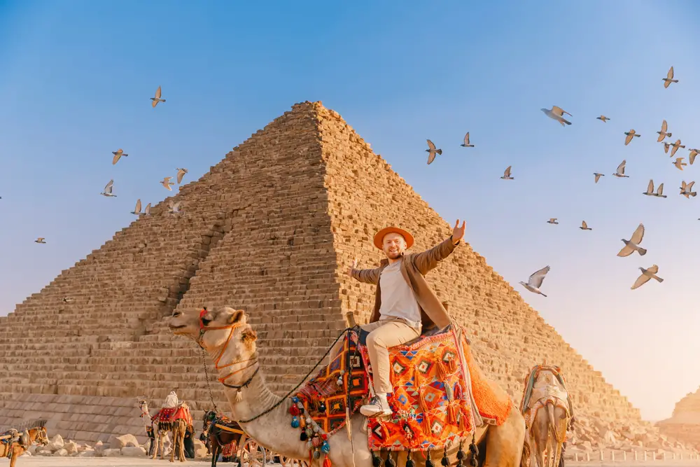 A man spreading her arms while riding a camel with birds flying in background and the Pyramid of Egypt. 