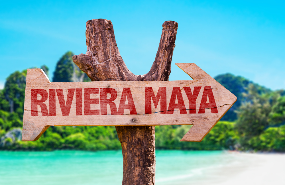 An wooden arrow signage saying Riviera Maya, and in background is a sea and mountains with trees.